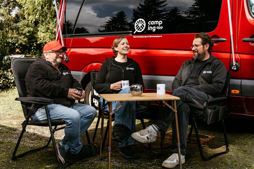 Jenny, Lukas & Dirk, das Team hinter der Camping App Social, sitzen fröhlich vor einem roten Camper