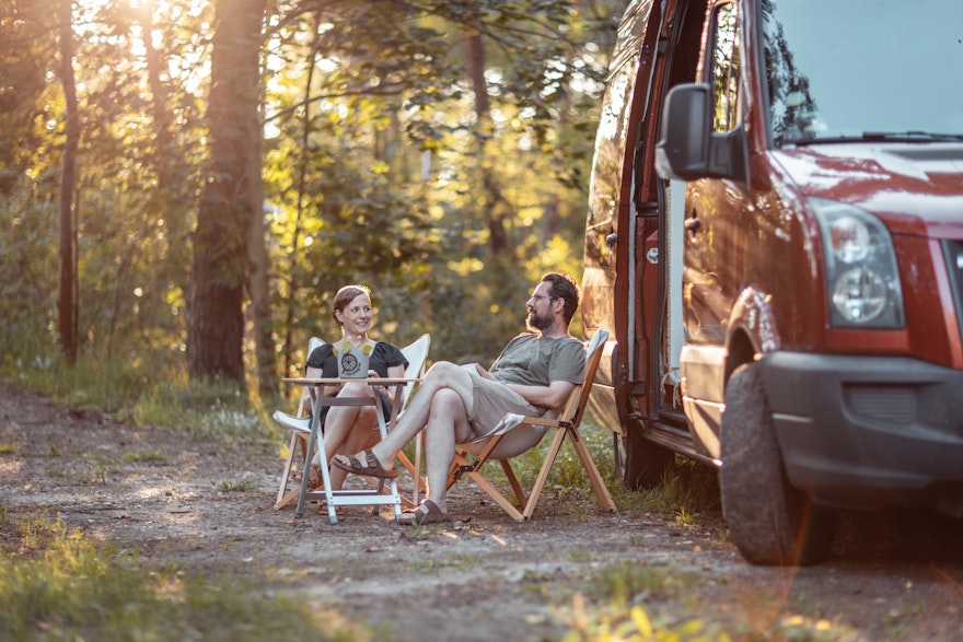 Die Macher der Camping App, Jenny & Lukas, sitzen vor ihrem roten Camper in einem idyllischen Wald und genießen die Abendsonne
