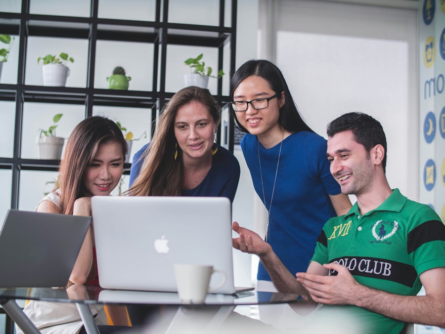 Four people looking at a white MacBook on a glass table