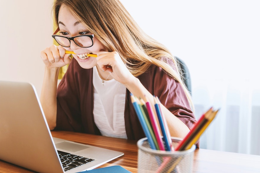 woman biting a pencil while sitting in a chair in front of a computer during the day