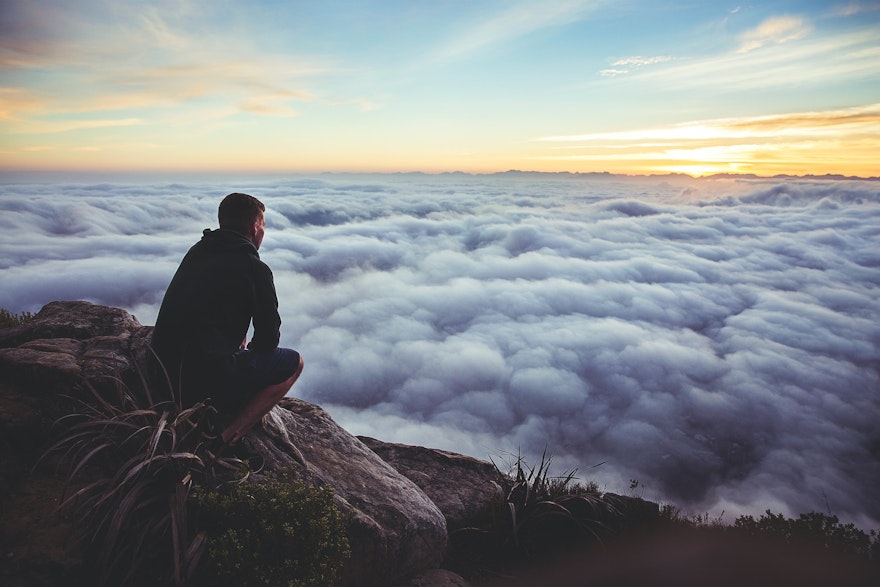A man squatting in a contemplative manner looking down at clouds from the top of a mountain. 