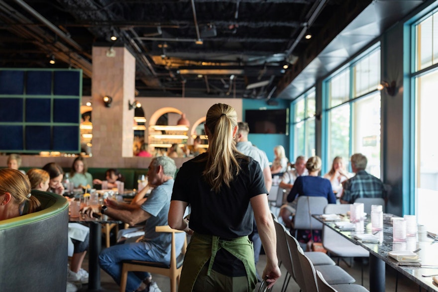 A waitress wearing a uniform serves a tray of food to a table in a bustling restaurant. The background is filled with other diners, tables, and staff, creating a lively and energetic atmosphere.