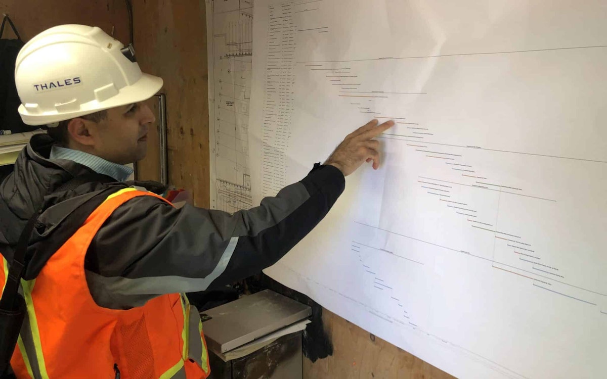 Construction supervisor wearing a hard hat and safety vest reviews and points to a project schedule posted on a job site wall.