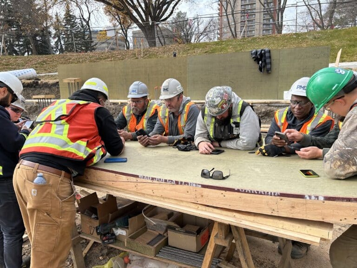 A group of construction workers wearing hard hats and high-visibility vests gather around a work surface at an active job site, checking information on their mobile phones. The image represents frontline collaboration, communication, and the use of simple digital tools to align crews on goals, progress, and productivity.