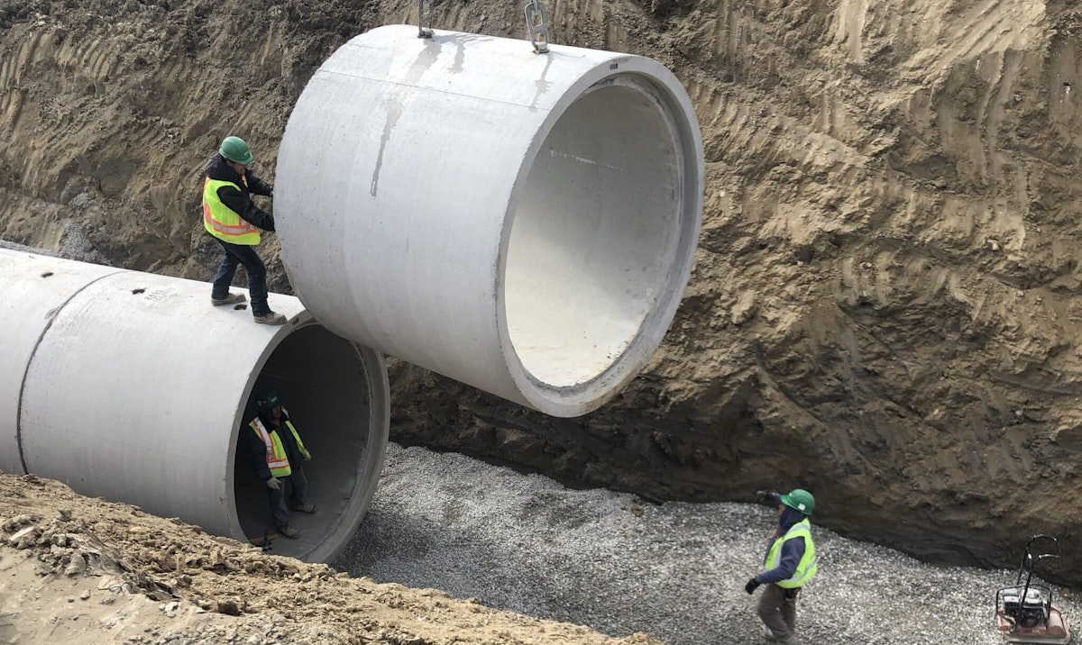 Construction workers install large precast concrete pipes in a trench, coordinating placement and alignment during a heavy civil project.