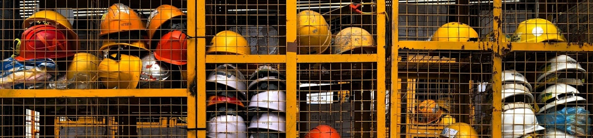 Construction hard hats of various colors stored in a metal cage on a job site, representing the construction workforce and frontline labor environment.