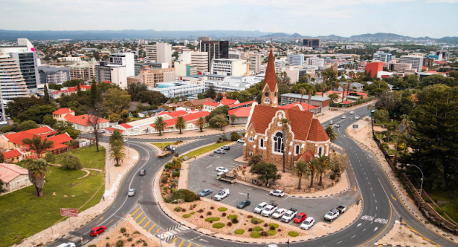 Photorealistic landscape image of Windhoek, Namibia, showcasing iconic landmarks like Christ Church, Independence Memorial Museum, and the Supreme Court of Namibia with a futuristic touch. The cityscape includes modern and colonial architecture, futuristic buildings, autonomous vehicles, and drones, set against rolling hills and desert landscape under a golden sunset.