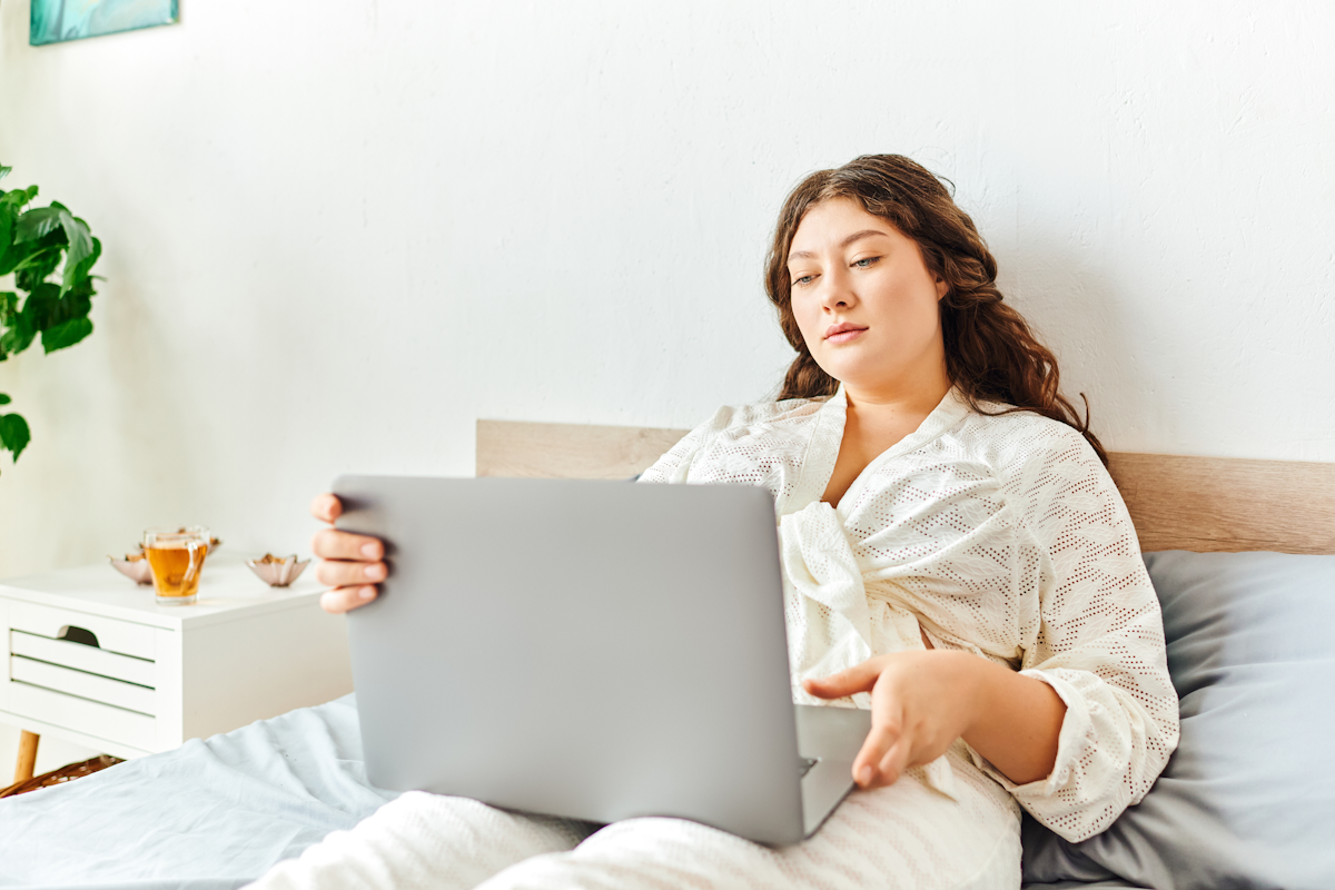 Woman sitting in bed with a laptop in soft natural light, representing reflection during a personal health journey.