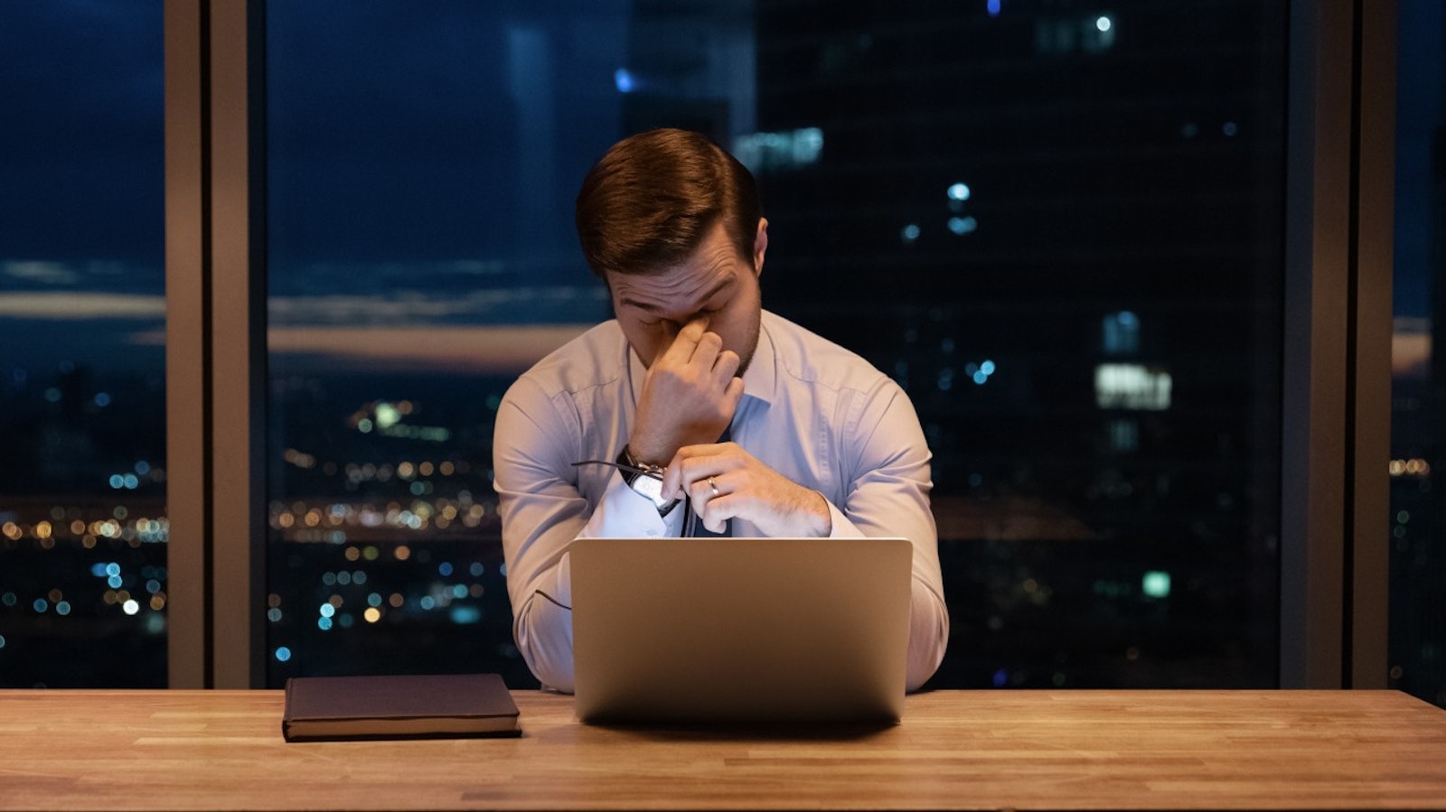 Stressed investment banker working late at night in a dimly lit office, surrounded by financial documents and a laptop, symbolizing the intense pressure of M&A deals.